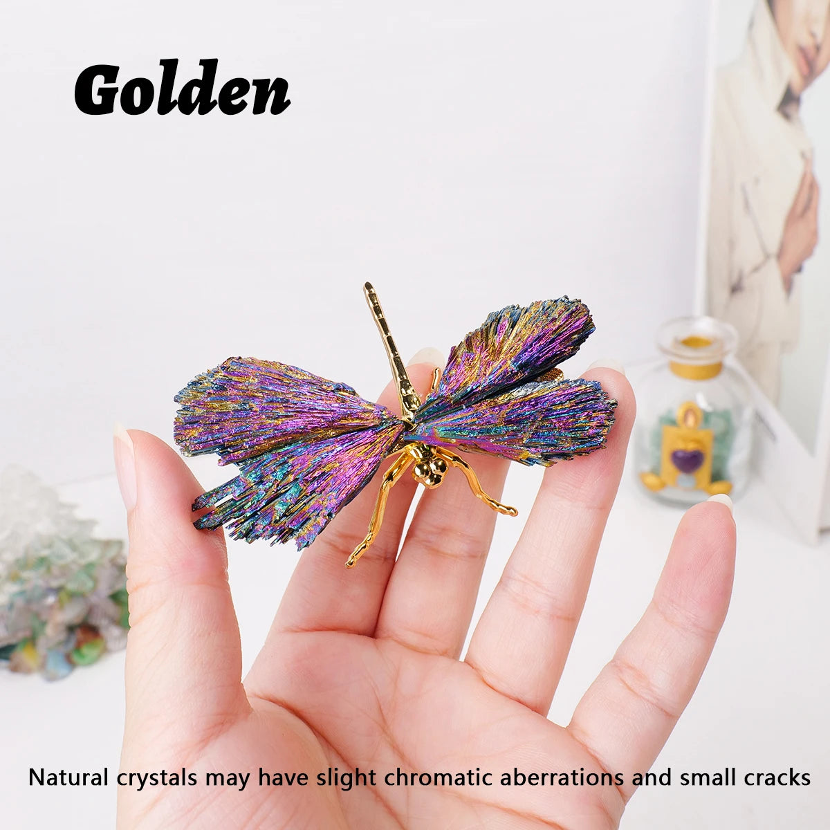 A hand holds a golden natural crystal tourmaline dragonfly figurine against a white background.