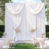 A white fabric backdrop with draped curtains and floral arrangements set up for an event.