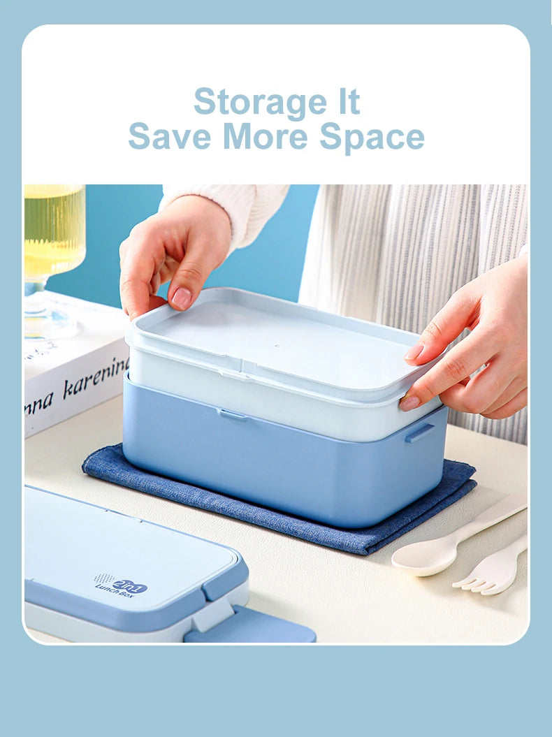 A light blue bento lunch box is shown open, with a hand placing it on a blue cloth next to a fork and spoon.