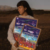 A person holds a framed print featuring Minecraft characters and landscapes outdoors.