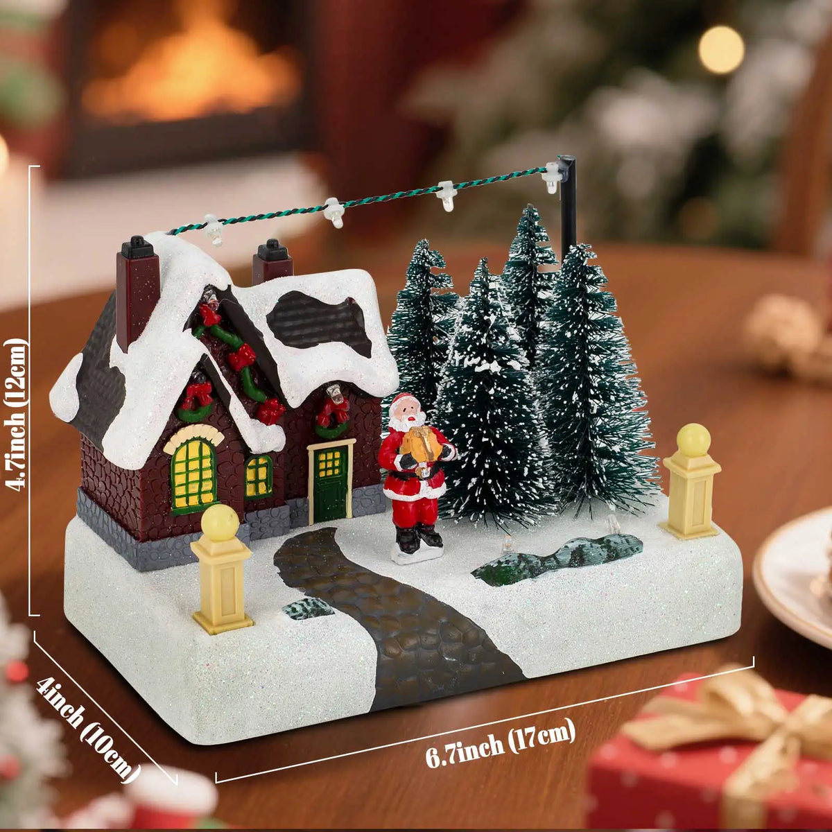 A miniature illuminated Christmas village scene featuring a brick house, Santa, and snowy evergreen trees on a white base.