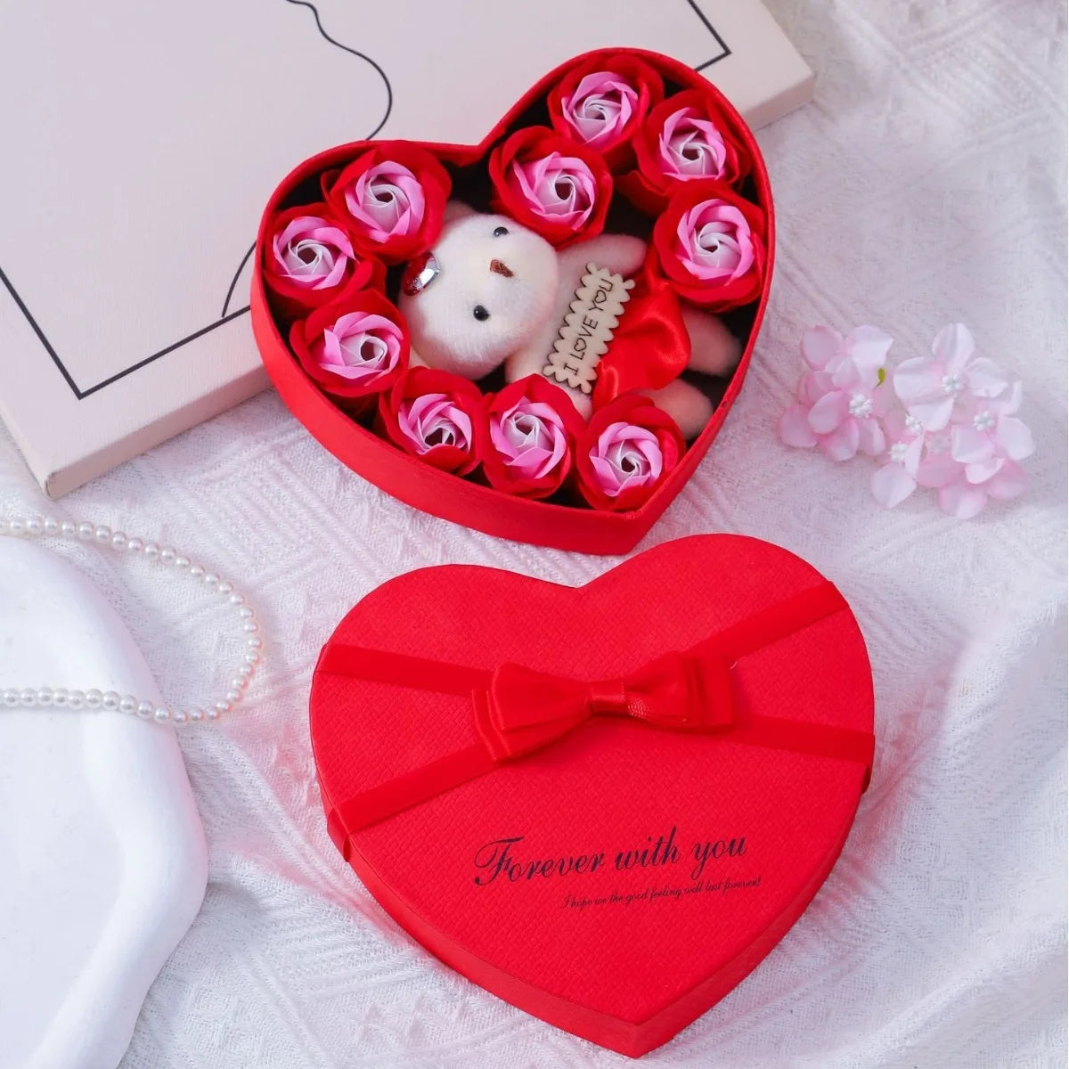 A red heart-shaped gift box filled with a white bear and pink rose flowers on a white textured surface.