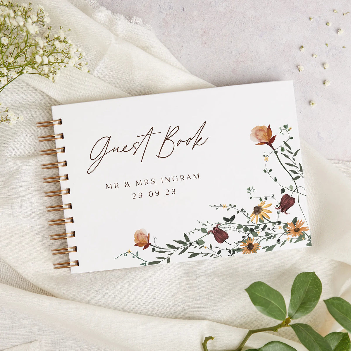A white personalized wedding guest book with gold binding and delicate floral watercolor accents on a cream linen background.