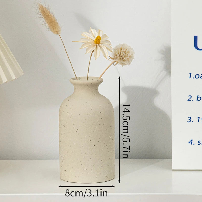 A minimalist ceramic vase with a speckled finish holds dried floral elements on a white shelf.