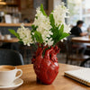 A red heart-shaped vase filled with white flowers sits on a wooden table.