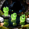 Green glow-in-the-dark resin charms of an alien cat and duck resting in a dark, mossy forest setting.