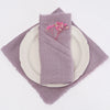 A stack of purple linen napkins with pink floral accents resting on a lavender cloth and a white plate.