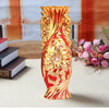 A tall, ornate gold-plated ceramic tabletop vase with a reddish-orange hue sits on a light surface.