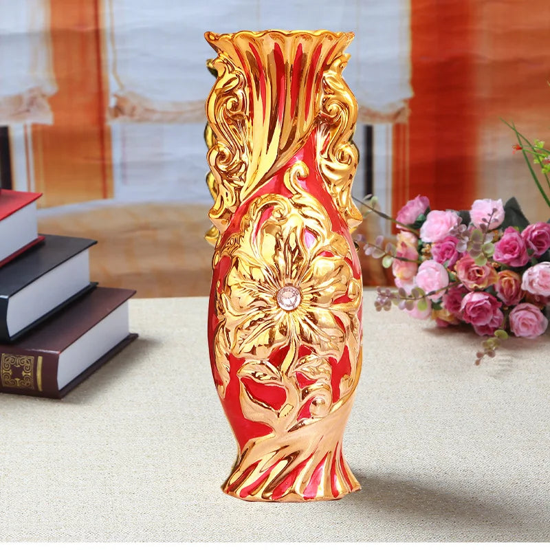 A tall, ornate gold-plated ceramic tabletop vase with a reddish-orange hue sits on a light surface.