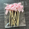 A set of pink bow cupcake toppers on wooden sticks packaged in a clear plastic bag.