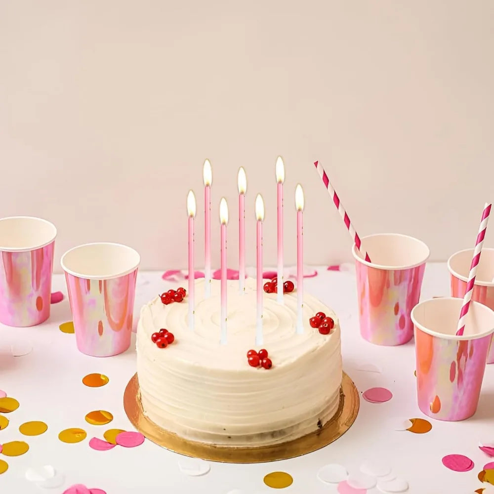 A round cake decorated with white frosting and lit birthday candles on a white table surrounded by pink cups.
