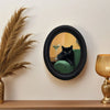 A framed painting of a black cat sitting on a green velvet cushion in an oval black frame.
