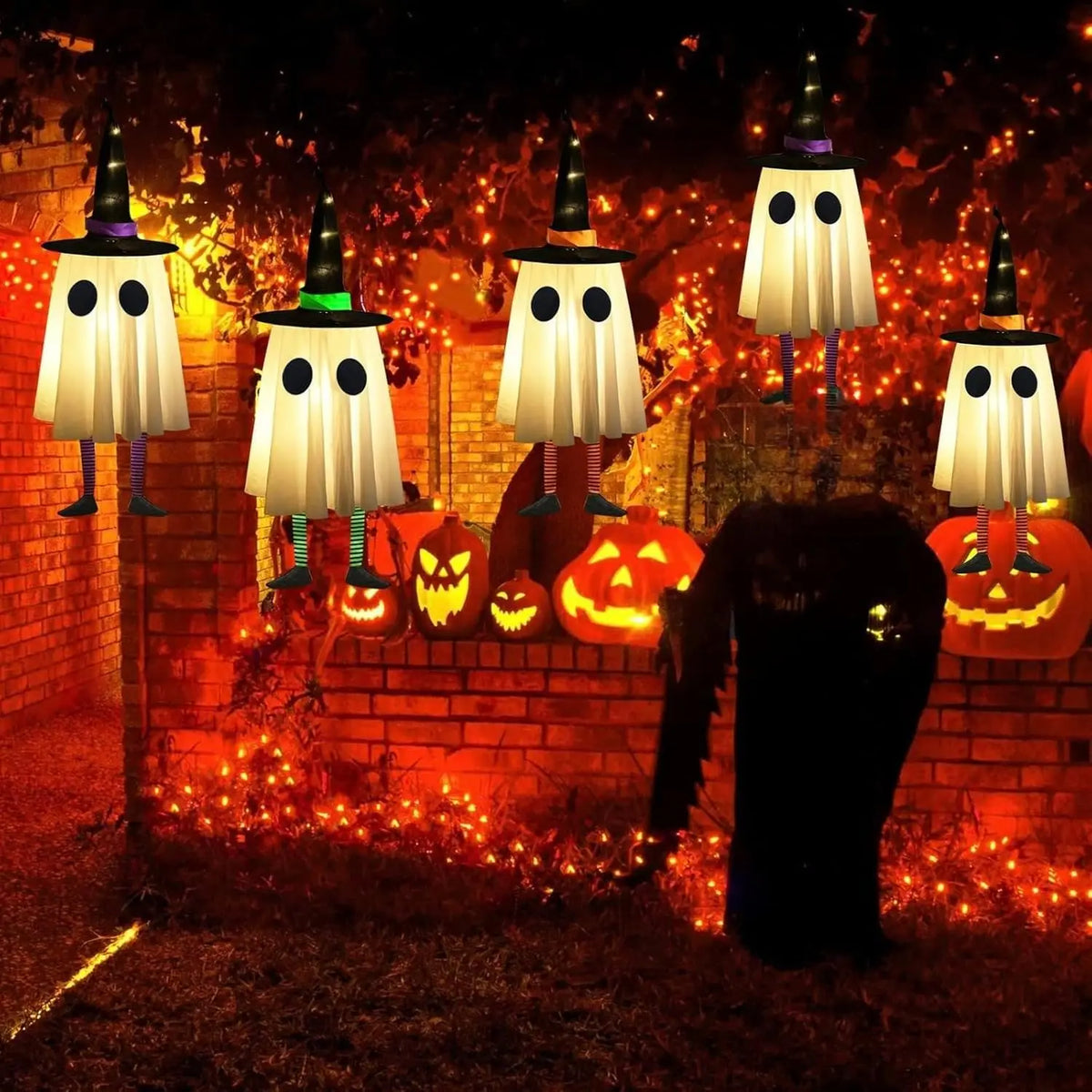 Halloween hanging ghost lights with witch hats illuminating a brick wall and orange pumpkins in a dark garden setting