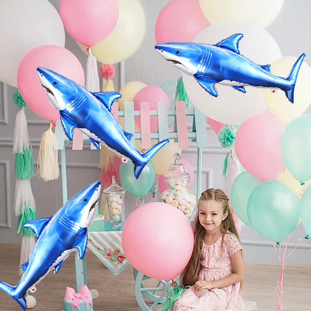 Blue shark foil balloons and a girl in a pink dress decorate an ocean-themed party setup.