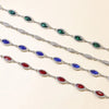 A display of colorful crystal waist chains featuring emerald green, sapphire blue, and ruby red gemstones.