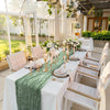 A long white table set with a light blue cheesecloth runner and floral decorations in an outdoor rustic setting.