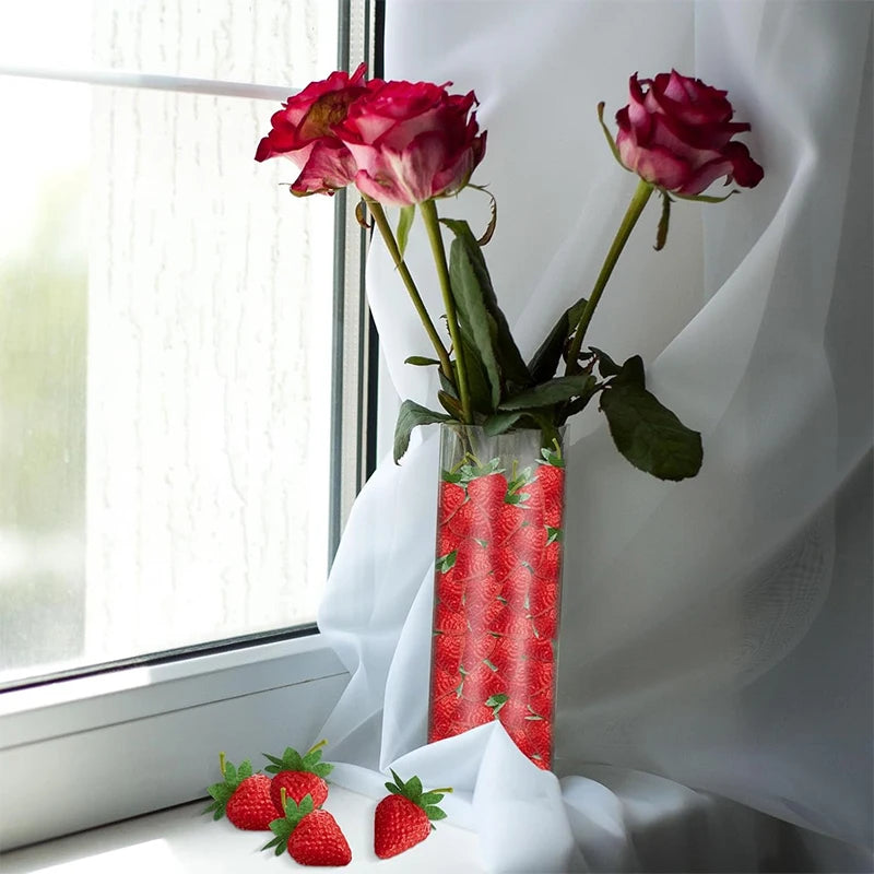 A glass vase holds several artificial red roses and fresh strawberries against a white curtain near a window.