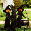 Three witch figures in black robes with orange carved faces sitting on green grass.