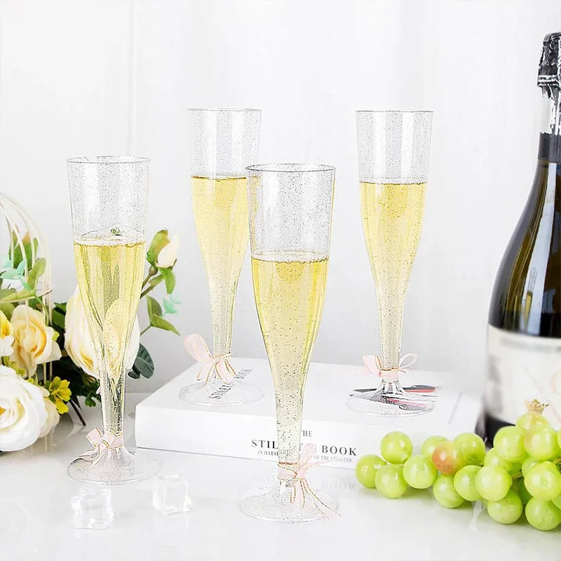 A set of glitter disposable champagne flutes displayed on a white surface with green grapes and a bottle.
