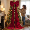 A woman in a sweater is decorating a Christmas tree with a large red velvet bow and long streamers.