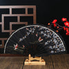 Elegant black folding fan decorated with white plum blossoms and gold accents on a wooden stand.