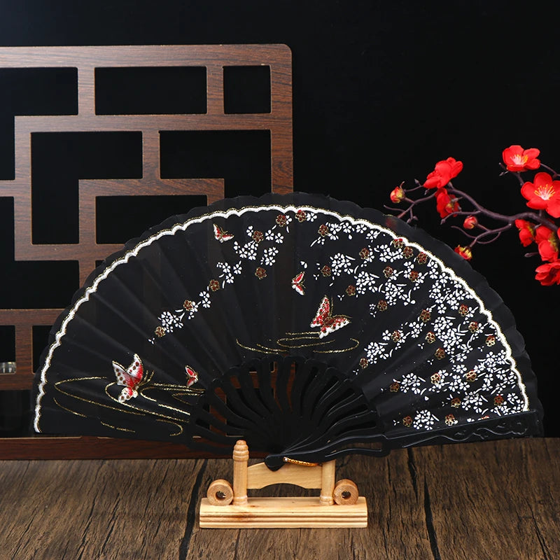 Elegant black folding fan decorated with white plum blossoms and gold accents on a wooden stand.