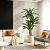 A tall artificial Dracaena tree in a white pot stands next to a modern sofa and a chair in a bright, minimalist living room.