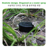 A black, disc-shaped device disguised as a water spray is partially buried in dirt and grass.