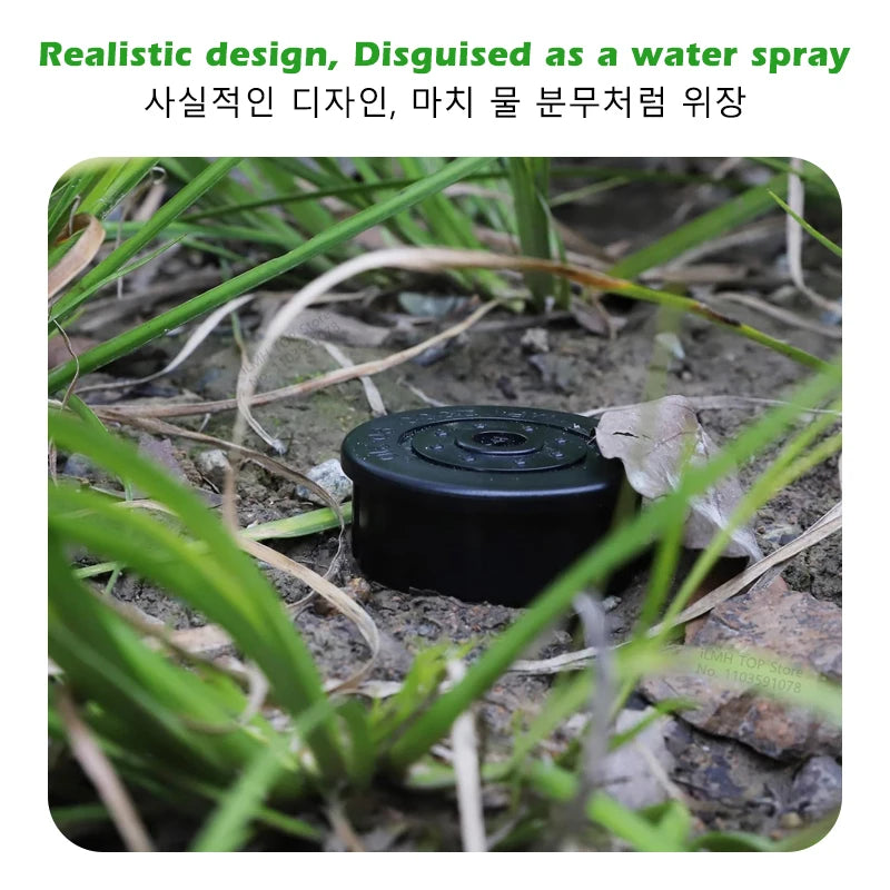 A black, disc-shaped device disguised as a water spray is partially buried in dirt and grass.