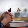 A hand holds an acrylic cross with a Bible verse inscribed on it, resting on an open book.