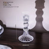 A clear glass pillar candle holder with an ornate design, shown against a dark background.