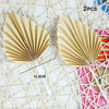 Two golden palm leaf cake toppers with flower decoration displayed on a white woven background.