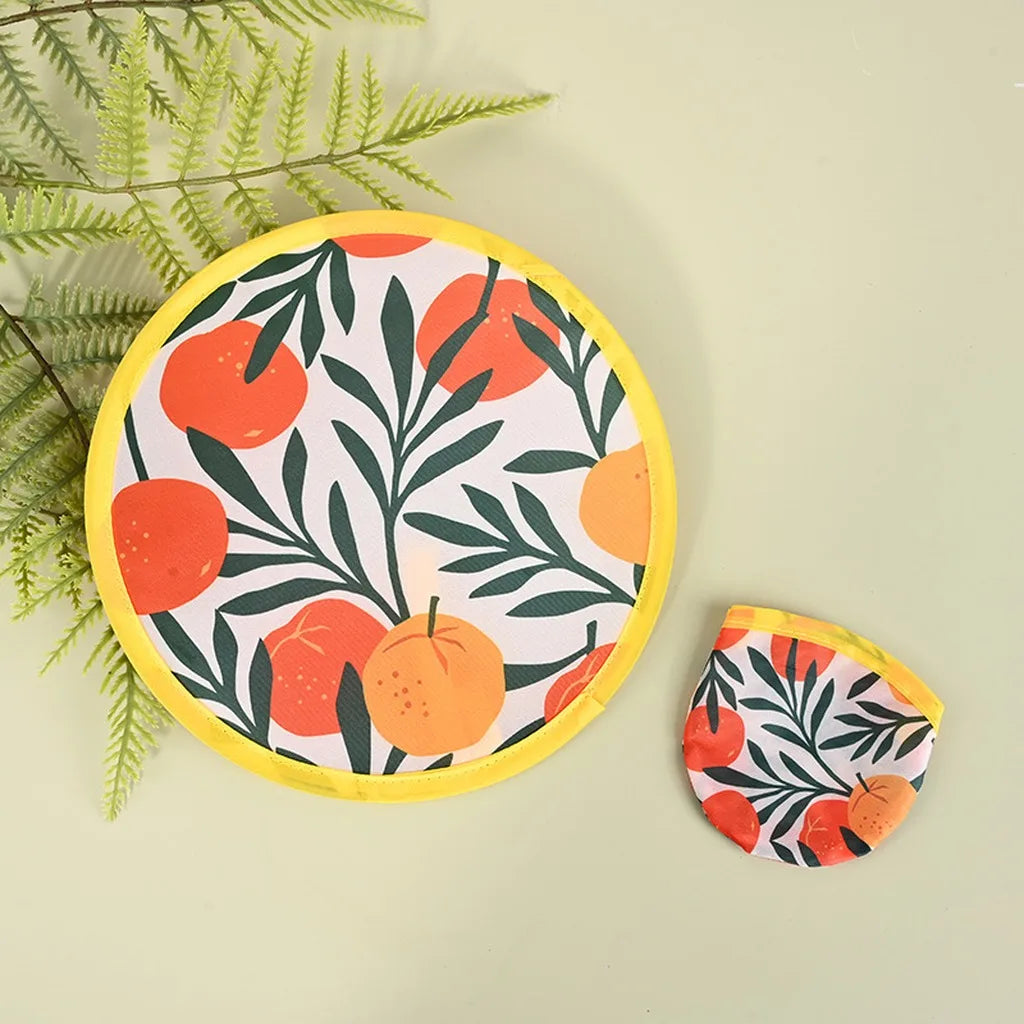A round fabric hand fan with a yellow border featuring a pattern of orange and green leaves on a light background.