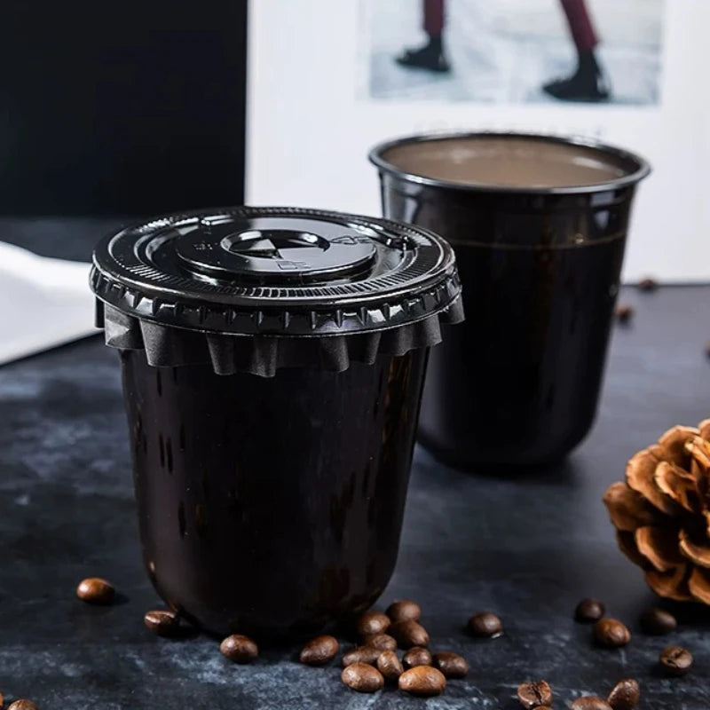 Eine schwarze Einwegtasse mit dunkler Flüssigkeit auf einer dunklen Oberfläche neben einer ähnlichen Tasse und Kaffeebohnen.