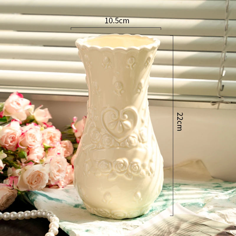 A white ceramic vase with embossed floral patterns is displayed on a surface next to pink roses.