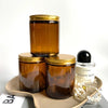 A set of two amber glass candle jars with gold metal lids resting on a light brown tray.
