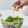 A hand is pouring a yellowish sauce from a small cup into a clear plastic bowl filled with green lettuce.