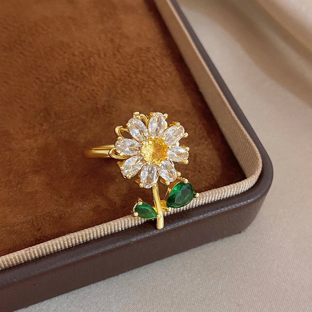 A gold cocktail ring featuring a white flower design with green leaves set in a brown velvet box.