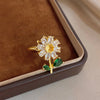 A gold cocktail ring featuring a white flower design with green leaves set in a brown velvet box.