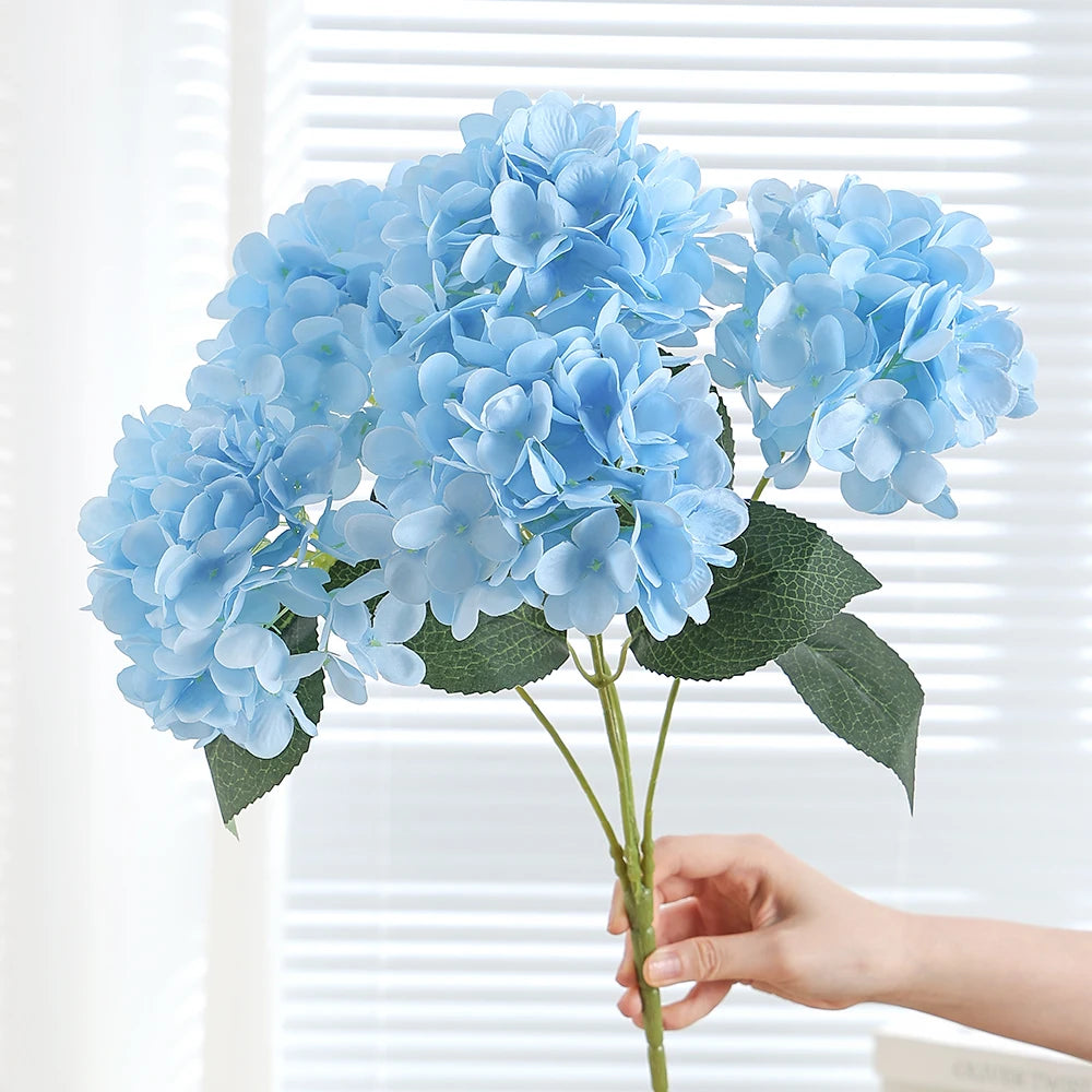 A bouquet of light blue artificial hydrangea silk flowers held by a hand against a white window blind.