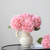 A cluster of pink artificial hydrangea flowers in a white vase on a black table.