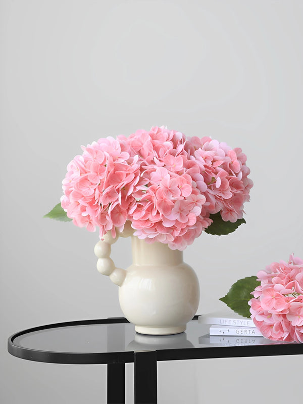 A cluster of pink artificial hydrangea flowers in a white vase on a black table.