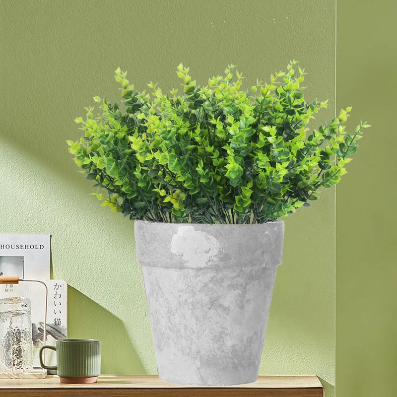 Plante d'eucalyptus artificielle en pot en céramique gris clair contre un mur vert pâle.