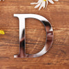 A shiny silver acrylic letter D cutout placed on a wooden surface with a small white flower nearby.