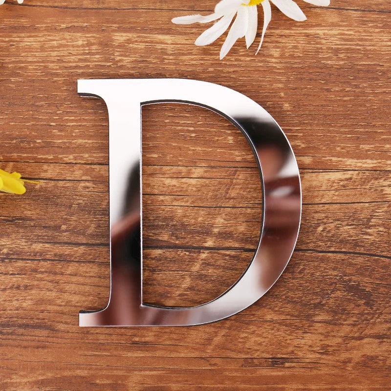A shiny silver acrylic letter D cutout placed on a wooden surface with a small white flower nearby.