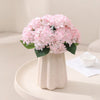 A bouquet of pink artificial hydrangea flowers in a white vase on a light surface.