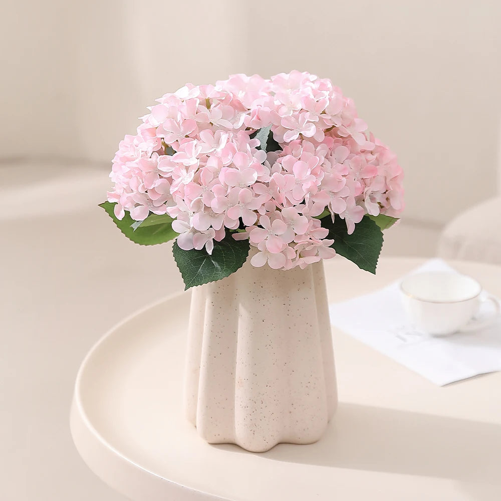 A bouquet of pink artificial hydrangea flowers in a white vase on a light surface.
