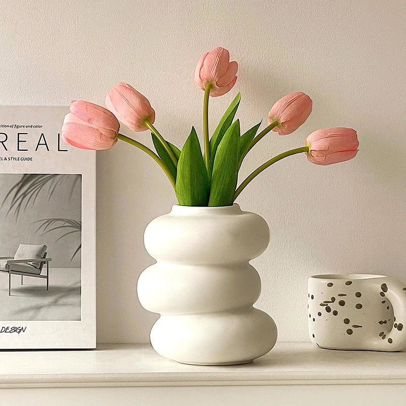 A vase of pink tulips in a white ceramic pot on a light shelf.