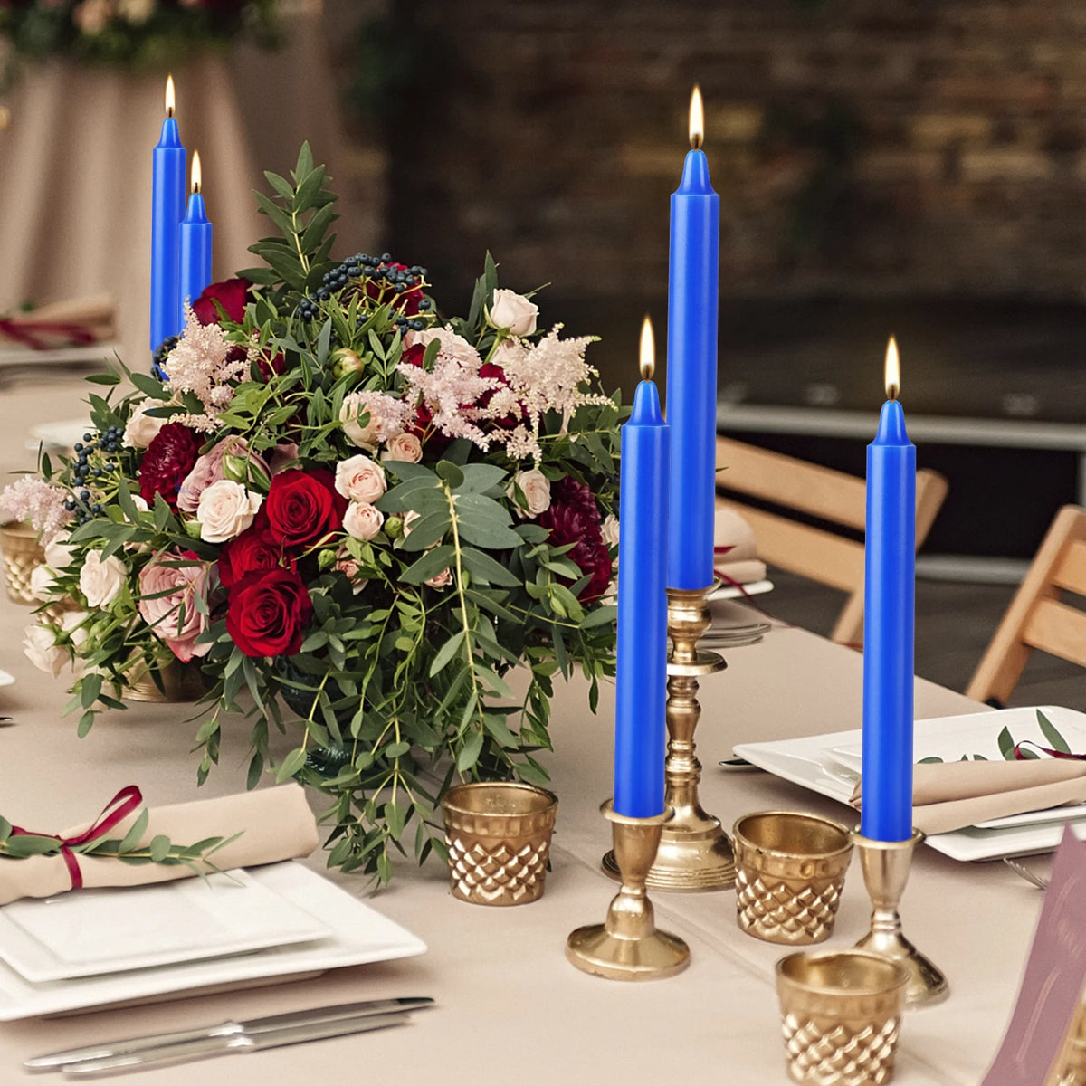 A floral arrangement of deep red and pink roses and greenery next to blue stick candles on a table.