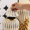 A hand is placing a gold-rimmed ceramic lid onto a white and gold striped decorative vase.